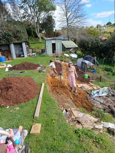 Eine Frau und ein Kind arbeiten in einem Garten und schaufeln Erde neben Mulchhaufen. Im Hintergrund sind Schuppen, Bäume und Häuser zu sehen. Ein Baby sitzt in einem Kinderwagen.