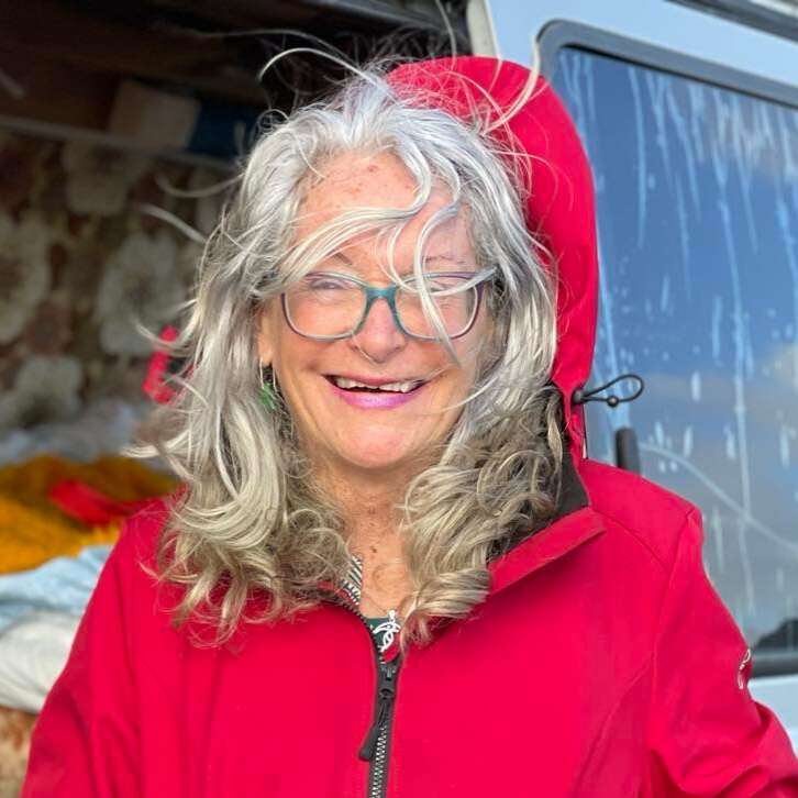 Das Bild zeigt eine ältere Frau mit langen, gewellten grauen Haaren, die eine rote Jacke mit Kapuze trägt und an einem windigen Tag vor einem weißen Lieferwagen steht.