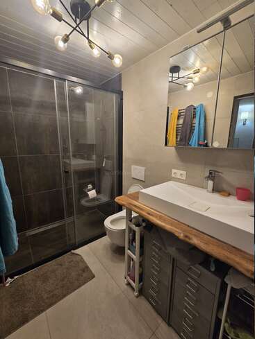Este moderno cuarto de baño cuenta con una cabina de ducha de cristal, un inodoro montado en la pared, una encimera de madera con un lavabo blanco, un gran espejo, un toallero y una elegante lámpara de techo.
