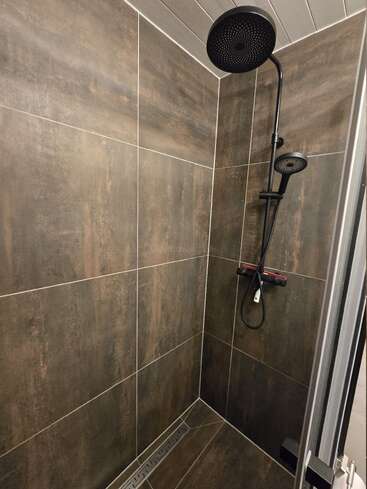 Esta moderna ducha cuenta con grandes azulejos oscuros similares a la piedra, una puerta de cristal, cabezal de ducha tipo lluvia y ducha de mano. El diseño es elegante, minimalista y contemporáneo, con líneas limpias.