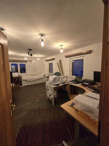 Una acogedora habitación con suelos de madera, varios escritorios, máquinas de coser, una cama, papeles esparcidos, ordenadores y bombillas colgantes expuestas, que crean un ambiente de espacio de trabajo creativo.