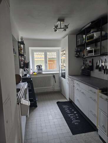 Se trata de una cocina estrecha y moderna con armarios blancos, encimeras grises y suelo de baldosas. Hay estantes en la pared, utensilios, electrodomésticos y una gran ventana. Ambiente acogedor.