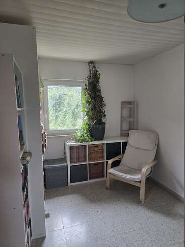 Una habitación pequeña y acogedora con una ventana, un sillón, una estantería, plantas, cubos de almacenaje y luz natural. Decoración minimalista, paredes claras y un ambiente tranquilo y acogedor.