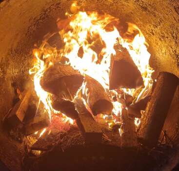 Big logs burn brightly in a contained fire pit. Orange flames dance and flicker, casting warm light against the soot-stained, circular metal surface.