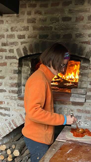 A person in an orange jacket prepares pizza, spreading tomato sauce on dough near a rustic brick oven with a wood fire burning brightly inside.