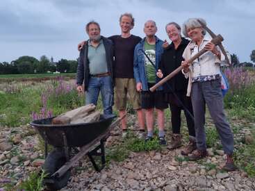 Five adults stand together smiling outdoors on rocky ground, next to a wheelbarrow filled with wood. One woman holds a pickaxe. Wildflowers grow nearby.