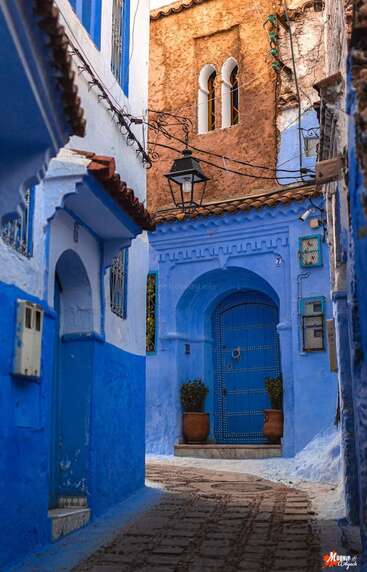 Dieses Bild zeigt eine schmale, verwinkelte Gasse in Chefchaouen, Marokko. Die Wände und Türen sind in leuchtendem Blau gestrichen und werden durch Töpferwaren, schmiedeeiserne Details und warmes Sonnenlicht hervorgehoben.