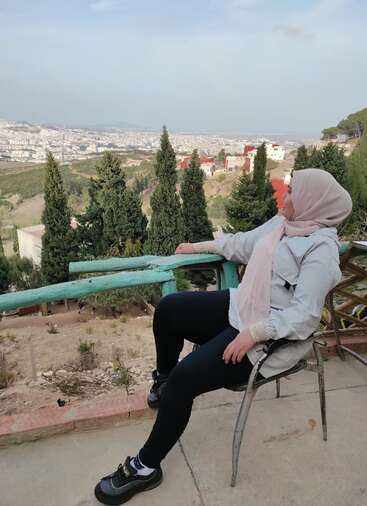 Eine Frau mit einem hellen Hijab sitzt auf einem Metallstuhl und schaut auf eine malerische Hügellandschaft mit Kiefern, Häusern und einer weitläufigen Stadt darunter.