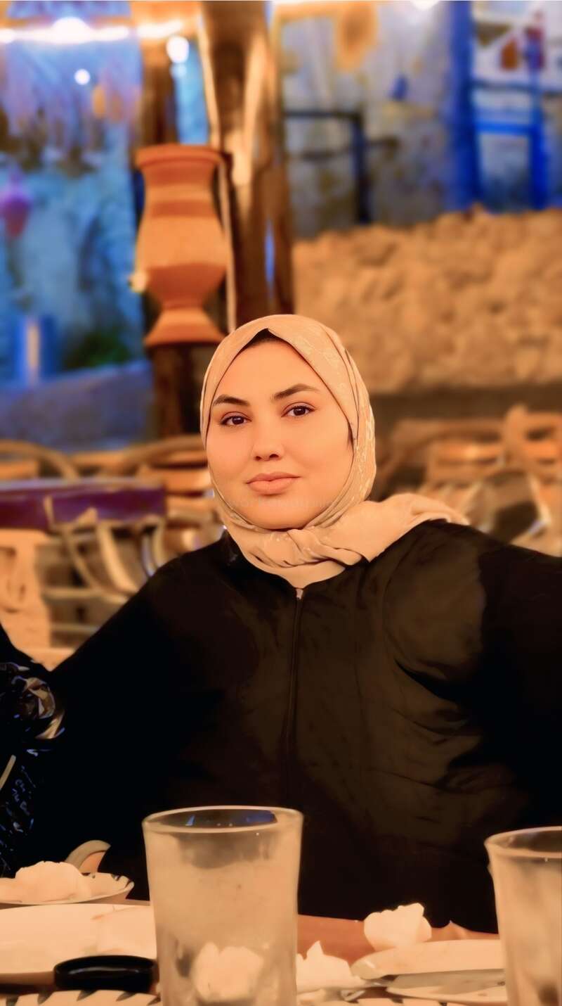 Eine Frau mit einem beigen Hijab sitzt an einem Restauranttisch. Der Hintergrund ist warm beleuchtet und gemütlich, was auf eine einladende und entspannte Atmosphäre beim Essen hindeutet.
