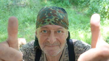 Um homem idoso sorridente, usando uma bandana e camisa de camuflagem, está ao ar livre, dando dois polegares para cima, com folhagem verde brilhante e luz do sol ao fundo.