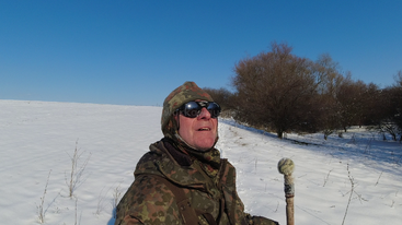 Um homem com equipamento de inverno camuflado e óculos de proteção está em um campo nevado, segurando uma bengala. Você pode ver as árvores nuas e o céu azul claro atrás dele.