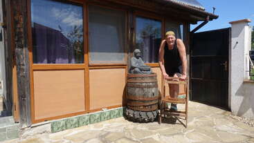 A person in a black tank top and shorts leans on a wooden chair outside a house, beside a Buddha statue sitting on a barrel and tire.