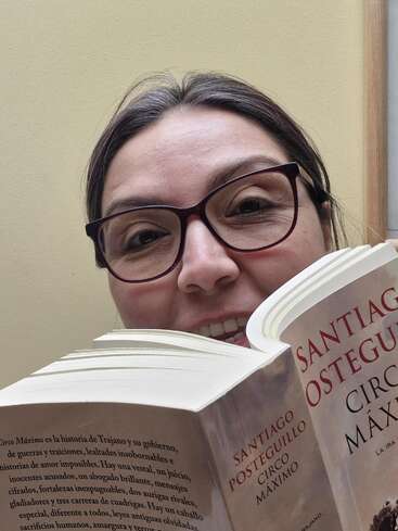 Uma mulher de óculos sorri enquanto segura e lê o livro "Circo Máximo", de Santiago Posteguillo. O plano de fundo é uma parede lisa de cor clara.