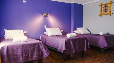 This cozy room features three single beds with purple bedspreads, white pillows, and rolled towels. The walls are purple and grey, with wooden flooring.