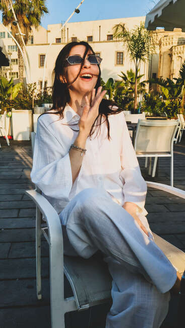Une femme vêtue de blanc est assise dehors, souriant joyeusement. Elle porte des lunettes de soleil, ses cheveux flottent au vent, elle profite d'une journée ensoleillée sur une terrasse. Atmosphère relaxante.