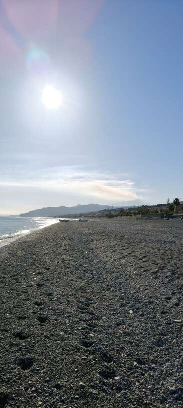 Un soleil éclatant brille sur une plage de galets aux vagues calmes, avec des montagnes lointaines et quelques personnes éparpillées profitant du paisible paysage côtier.