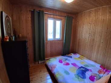 This cozy wooden room features a colorful bedspread, small dresser with decorations, two green curtains framing a window, and warm, inviting wood-paneled walls and ceiling.