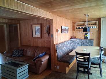 This cozy wooden cabin interior features a brown leather sofa, corner dining area with patterned benches, a light table, shelves with jars, artwork, and warm lighting.