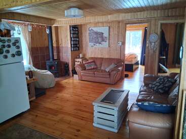 This cozy cabin living room features wood-paneled walls, leather sofas, a wood stove, rustic decor, natural light, a white fridge, and visible bedroom entrances.