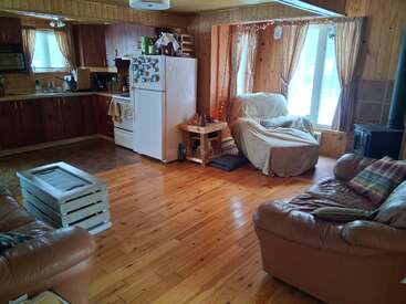This cozy living room features wood-paneled walls, two leather sofas, a covered armchair, a rustic kitchen, large windows with curtains, and a white refrigerator.