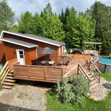 A cozy wooden house with a spacious deck, outdoor seating, umbrella-covered table, above-ground pool, surrounded by lush green trees and natural landscape, inviting relaxation.