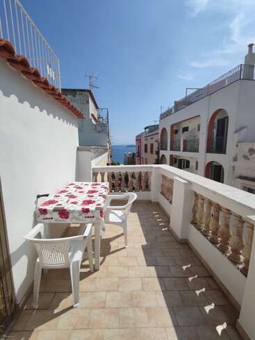 Sonniger Balkon mit gefliestem Boden, weißem Geländer, Plastikstühlen und einem mit Blumentuch bedeckten Tisch. Mit Blick auf die Nachbargebäude und in der Ferne auf das glitzernde Meer.