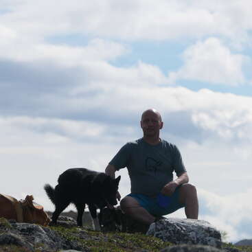 Un hombre se arrodilla en un terreno rocoso junto a un perro negro. Sostiene una taza azul. El cielo está cubierto de nubes dramáticas, creando una escena tranquila al aire libre.
