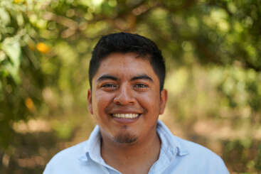 Un homme souriant se tient à l'extérieur, vêtu d'une chemise bleu clair. L'arrière-plan est composé de verdure floue et de lumière du soleil, créant une atmosphère chaleureuse, joyeuse et naturelle.
