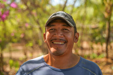 Un homme moustachu sourit chaleureusement à la caméra, vêtu d'une casquette et d'une chemise bleue. L'arrière-plan est en plein air, avec du feuillage vert et la lumière du soleil.