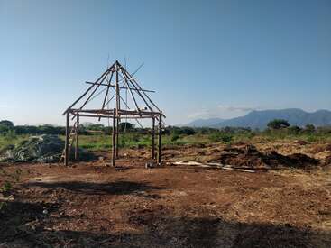 Une simple structure en bois en cours de construction se dresse sur un sol nu, entouré de champs verts et de montagnes lointaines, sous un ciel bleu clair, par une journée ensoleillée.