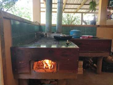 Cette image montre une cuisine rustique en plein air avec un poêle à bois. Un feu brûle à l'intérieur et des casseroles, une bouilloire et des ustensiles de cuisine sont posés dessus.