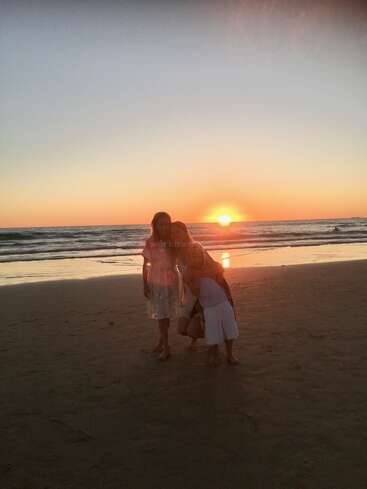 Three people pose together on the sandy beach during a beautiful sunset, with waves in the background and the sky painted with warm orange hues.