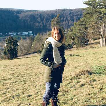 A woman stands smiling on a grassy hillside, dressed warmly in a green coat, scarf, and boots. Behind her are trees, distant hills, and buildings.