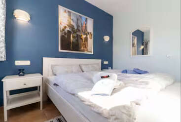 This cozy bedroom features a large white bed, blue accent wall, framed artwork, nightstand with lamp, soft lighting, mirror, and neatly folded towels on the bed.