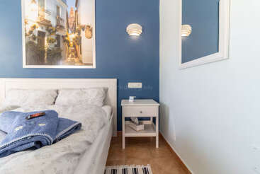 A cozy bedroom features a white bed with patterned bedding, a blue wall, framed artwork, nightstand, lamp, mirror, and neatly folded blue towels with a remote.