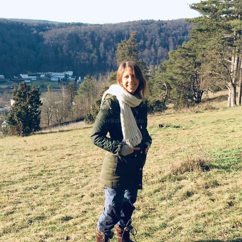 A woman stands smiling on a grassy hillside, dressed warmly in a green coat, scarf, and boots. Behind her are trees, distant hills, and buildings.