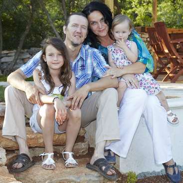 La imagen muestra a una familia de cuatro miembros sentados en unos escalones de piedra, con los padres abrazando a sus dos hijas pequeñas, todos vestidos con ropa informal y sandalias.