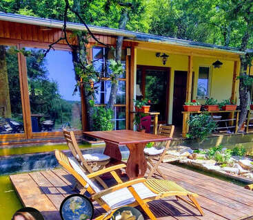 Eine gemütliche Holzterrasse mit Tisch und Stühlen bietet einen Blick auf einen ruhigen Teich und grenzt an ein charmantes Haus, das von üppigen Bäumen und sattgrünen Pflanzen umgeben ist.