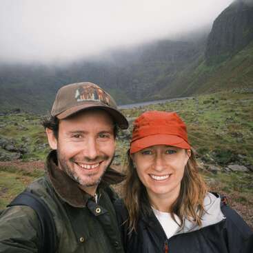 Ein lächelndes Paar mit Mützen und Outdoor-Jacken posiert für ein Selfie in einer üppigen, nebligen Berglandschaft mit felsigem Gelände und nebligen Hügeln hinter ihnen.