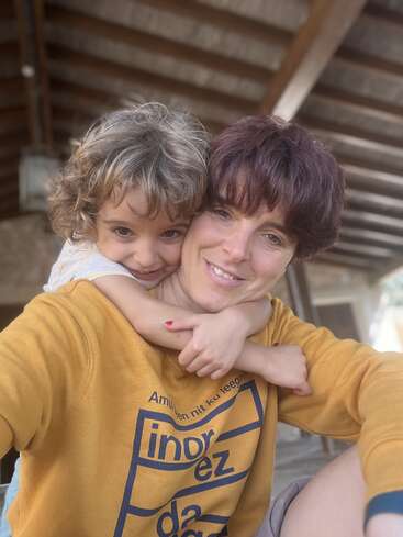 Uma mulher e uma criança pequena estão sorrindo, compartilhando um abraço afetuoso. Eles parecem felizes, desfrutando de um momento de ternura juntos sob uma estrutura de telhado de madeira.