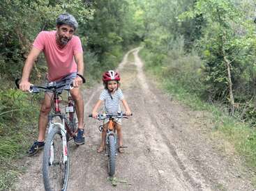 Um homem e uma criança estão andando de bicicleta em uma trilha de terra em uma floresta. A criança usa um capacete vermelho, e ambos parecem felizes e ao ar livre.
