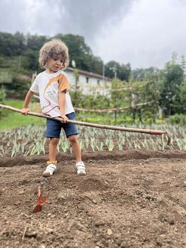 Uma criança de cabelos cacheados usa uma enxada de cabo longo em um jardim. O céu está nublado, e os vegetais crescem em fileiras bem cuidadas atrás deles.