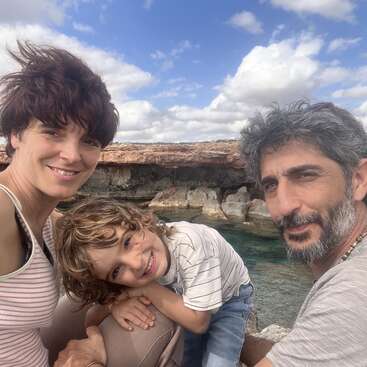 Uma família feliz de três pessoas posa ao ar livre perto da água e de penhascos rochosos. Eles sorriem calorosamente, curtindo um dia ensolarado juntos sob um céu azul parcialmente nublado.