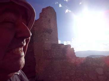 Das Bild zeigt das Profil eines Mannes, der einen Hut trägt, vor einer Steinmauer und einer hellen Sonne, die am Himmel mit einigen Wolken scheint.