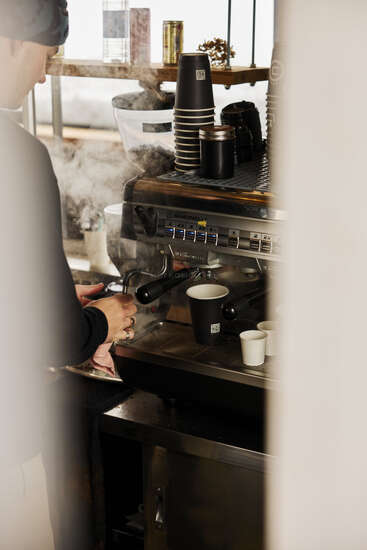 Un barista prépare du lait à la machine à café, entouré de tasses en papier, la vapeur s'élevant, dans un café confortable à la lumière naturelle douce et à l'atmosphère chaleureuse.