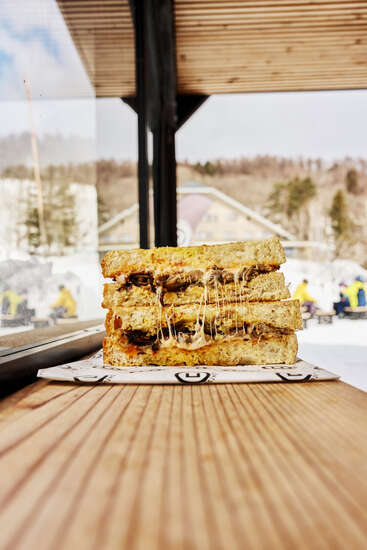 Un délicieux sandwich au fromage grillé, au fromage fondant et à la garniture consistante, est posé sur une table en bois, avec une scène extérieure enneigée en arrière-plan.