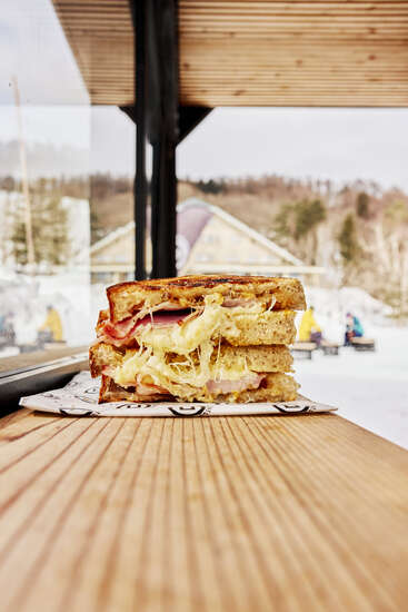 Un délicieux sandwich au fromage grillé et au jambon est posé sur une table en bois, empilé et débordant de fromage fondu. On aperçoit une scène hivernale en plein air.