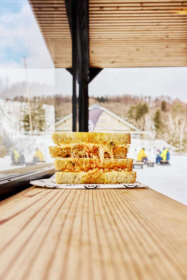 Un délicieux sandwich au fromage grillé est posé sur une table en bois. L'arrière-plan montre une scène extérieure enneigée avec des personnes portant des vêtements d'hiver colorés. Une ambiance chaleureuse.