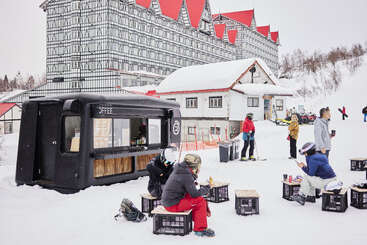 Un petit kiosque à café noir sert des personnes assises sur des caisses dans la neige, à proximité d'un grand hôtel aux toits rouges et de skieurs enthousiastes.