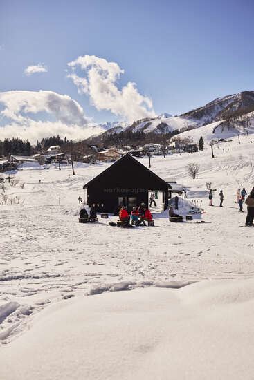 Des personnes se rassemblent à l'extérieur d'une petite cabane noire dans un décor de montagne enneigée, entourée de maisons pittoresques, d'arbres et d'un ciel bleu vif avec des nuages épars.